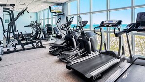 Un gimnasio con cintas de correr, bicicletas estáticas y máquinas de pesas, amplias ventanas con vistas a la playa.