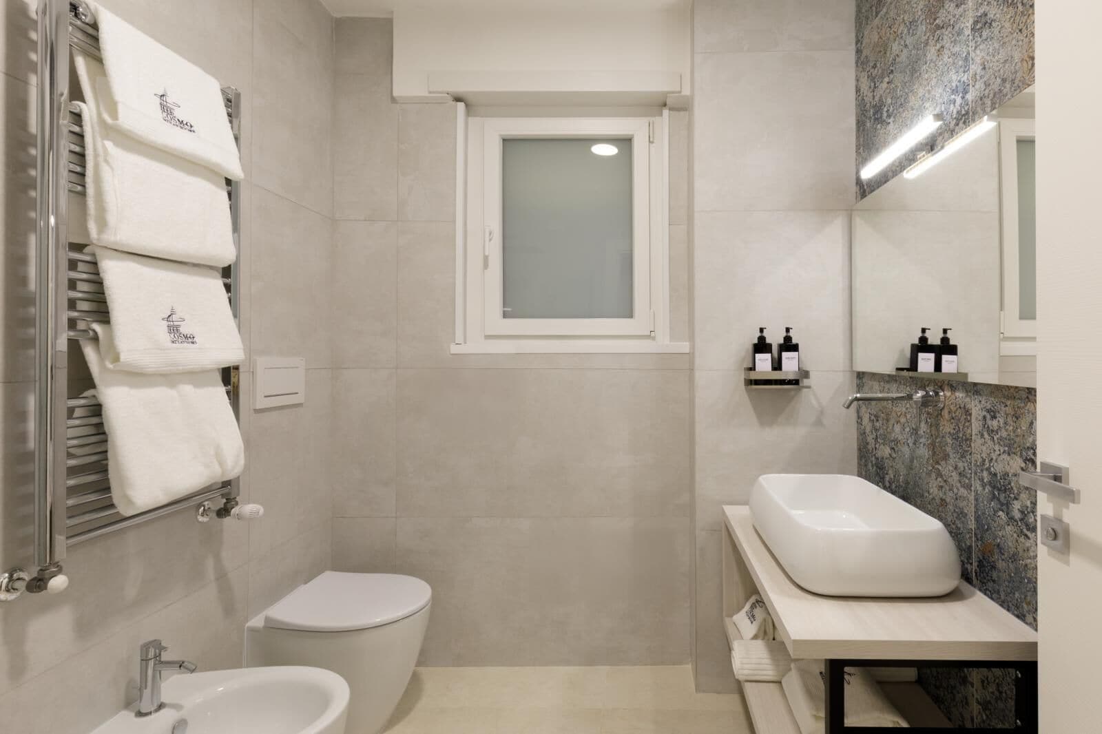 A modern bathroom with a toilet, bidet, and sink. There are towels hanging on a rack and a window above the toilet.