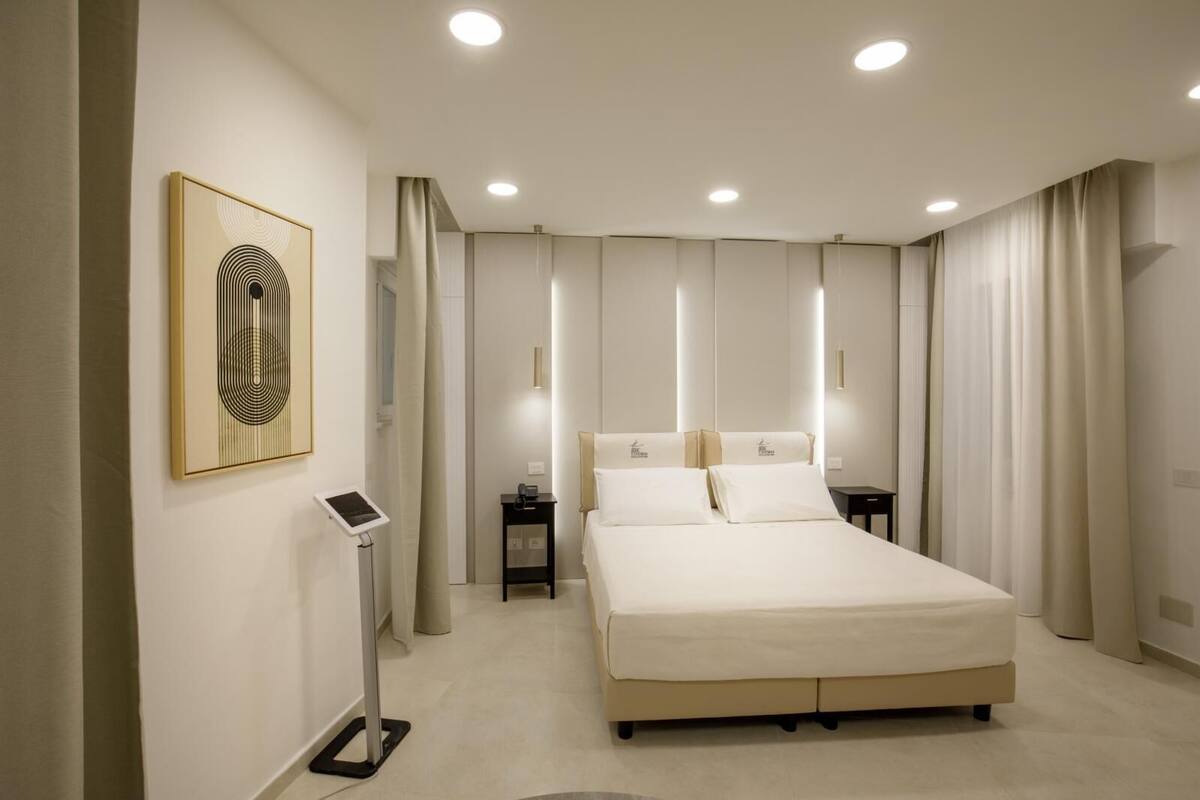 A modern hotel room with a large bed, bedside tables, a digital device, and a framed artwork on the wall.