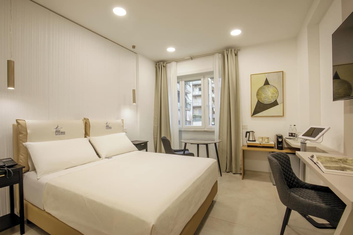 A hotel room with a bed, a desk, a chair, a window with curtains, and a painting on the wall.