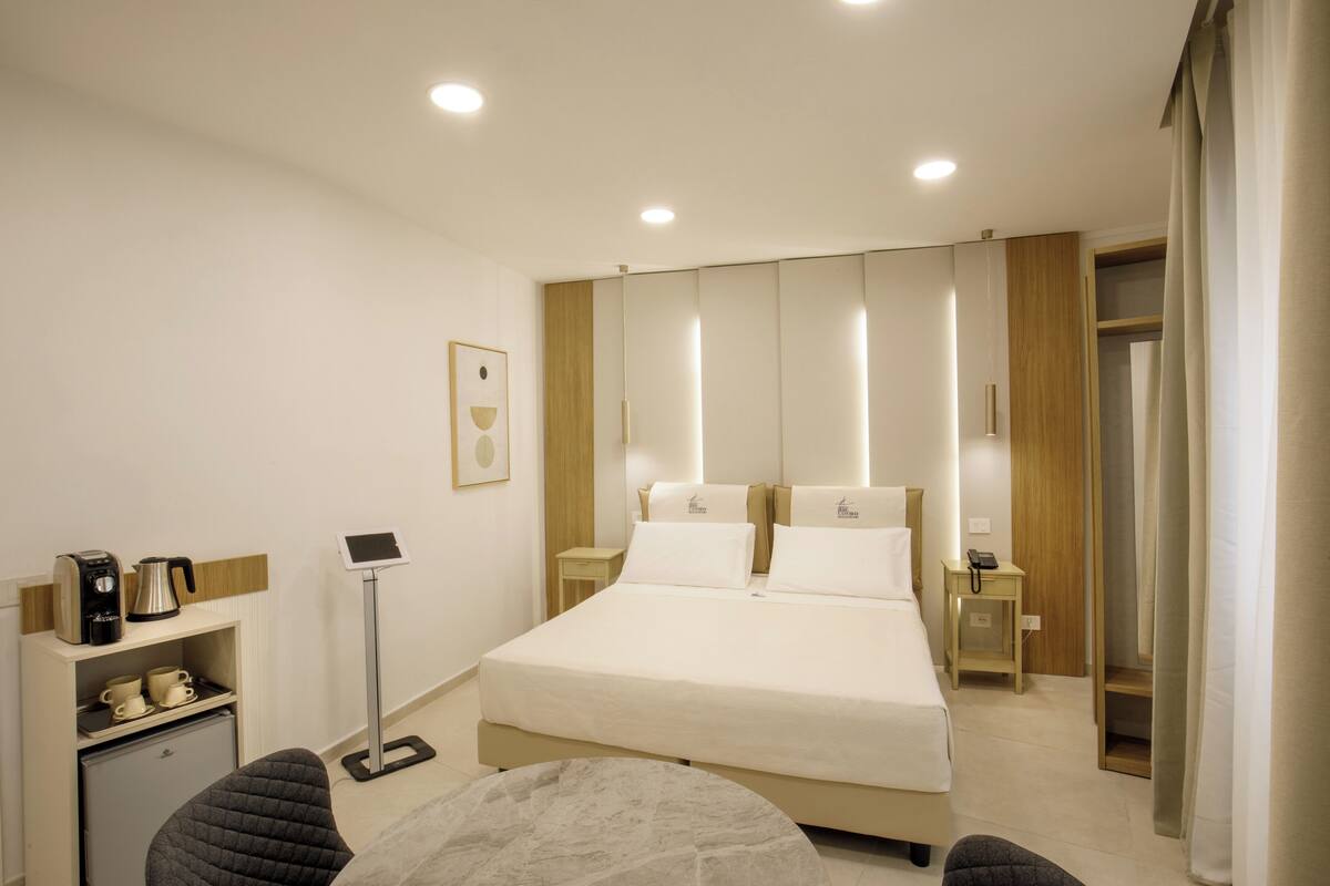 A hotel room with a bed, bedside tables, a desk with a chair, a coffee maker, and a small refrigerator.