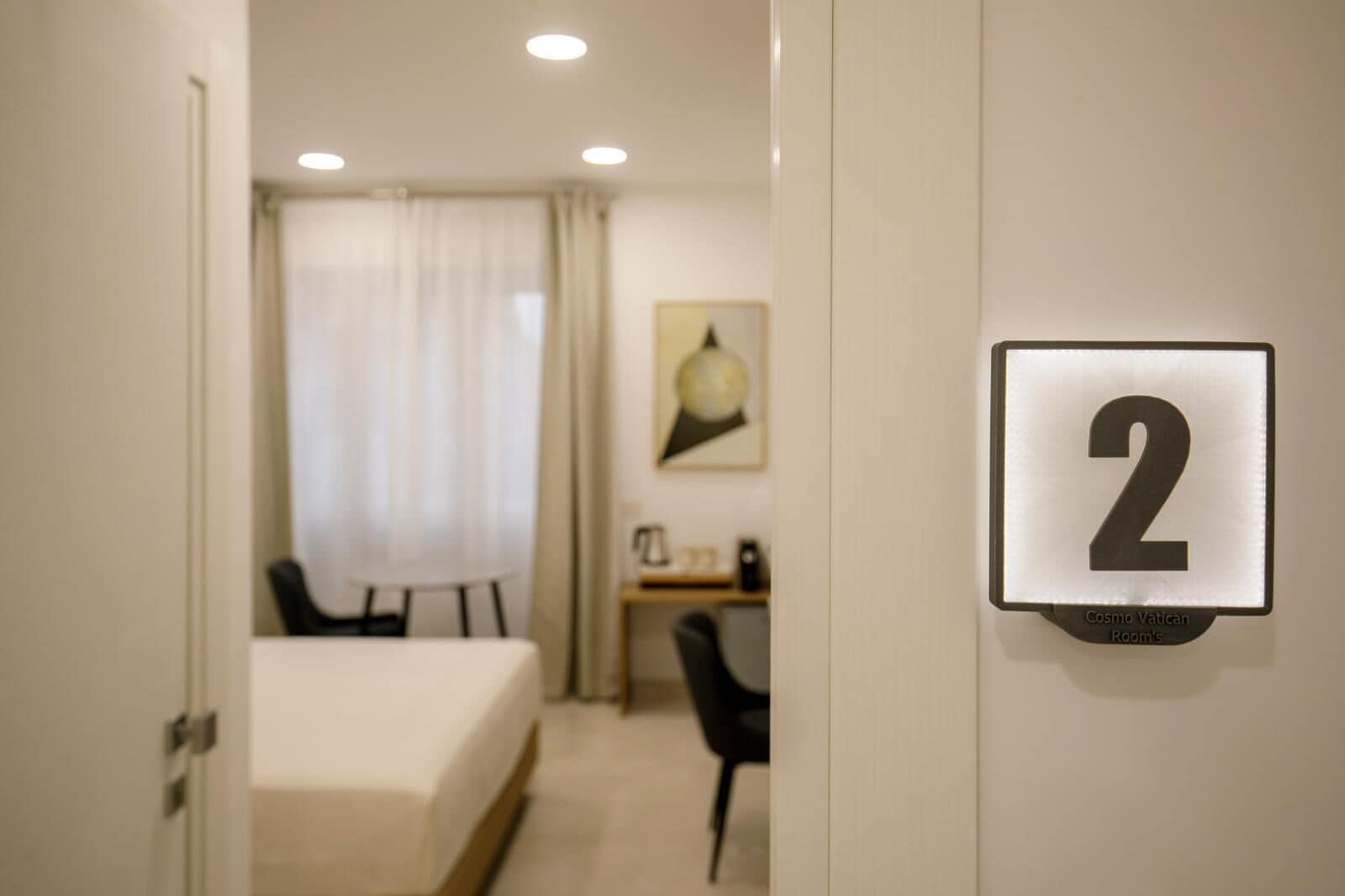 Ein Hotelzimmer mit einem Bett, einem Schreibtisch und einem Stuhl. Ein Schild zeigt die Zimmernummer 2 an.