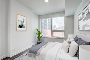 A modern bedroom with a large window, a bed with multiple pillows, a bedside table, and a framed picture on the wall. - Spacious Modern 2-Bedroom in Calgary | BLVD Beltline (Calgary)