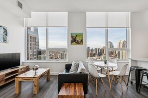 Ein modernes Wohnzimmer mit großem Fernseher, Holzmöbeln, einem schwarzen Sofa, weißen Sesseln und Blick auf die Stadt durch große Fenster.