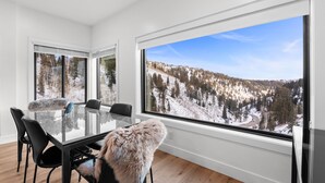 Un comedor moderno con una mesa y sillas de vidrio, ventanas amplias con vistas a una montaña nevada y un cubrepuesto de piel en una silla.