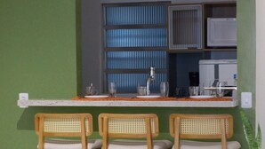 Ein Essbereich mit einem weißen Tisch und sechs Holzstühlen mit gewobenen Rückenlehnen. Im Hintergrund ist eine kleine Küchenzeile mit einer Mikrowelle und einem Kühlschrank erkennbar.