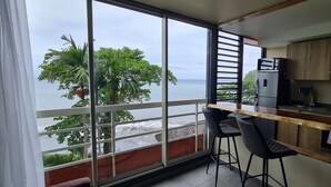 A balcony with a view of the sea, a wooden bar counter, two black bar stools, and a refrigerator.