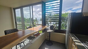 A small kitchen with a wooden table and chairs, a refrigerator, and a window with a view of palm trees and a body of water.