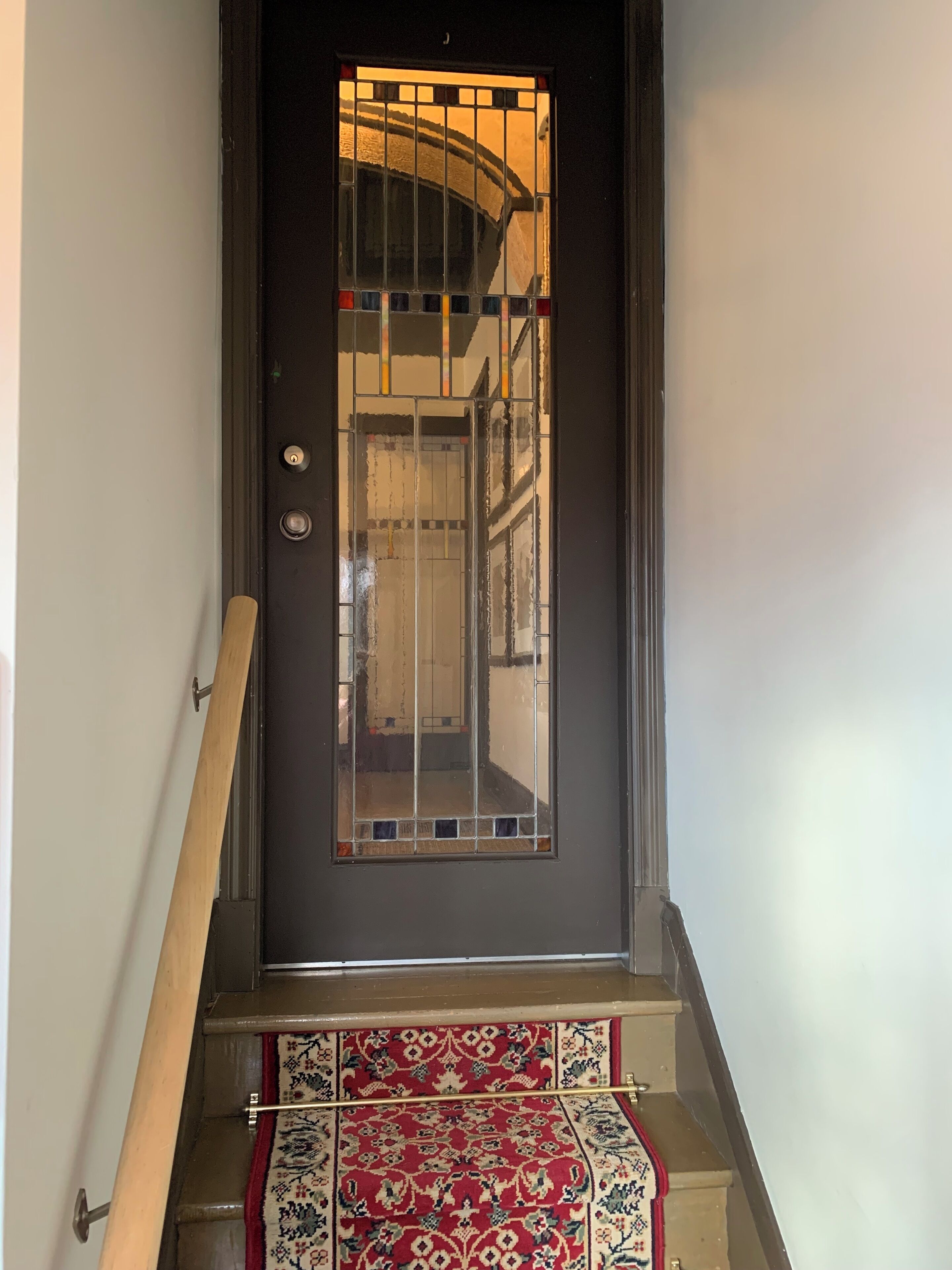 Puerta oscura con un panel de vidrio y una reja metálica, que da acceso a una escalera con alfombra estampada.