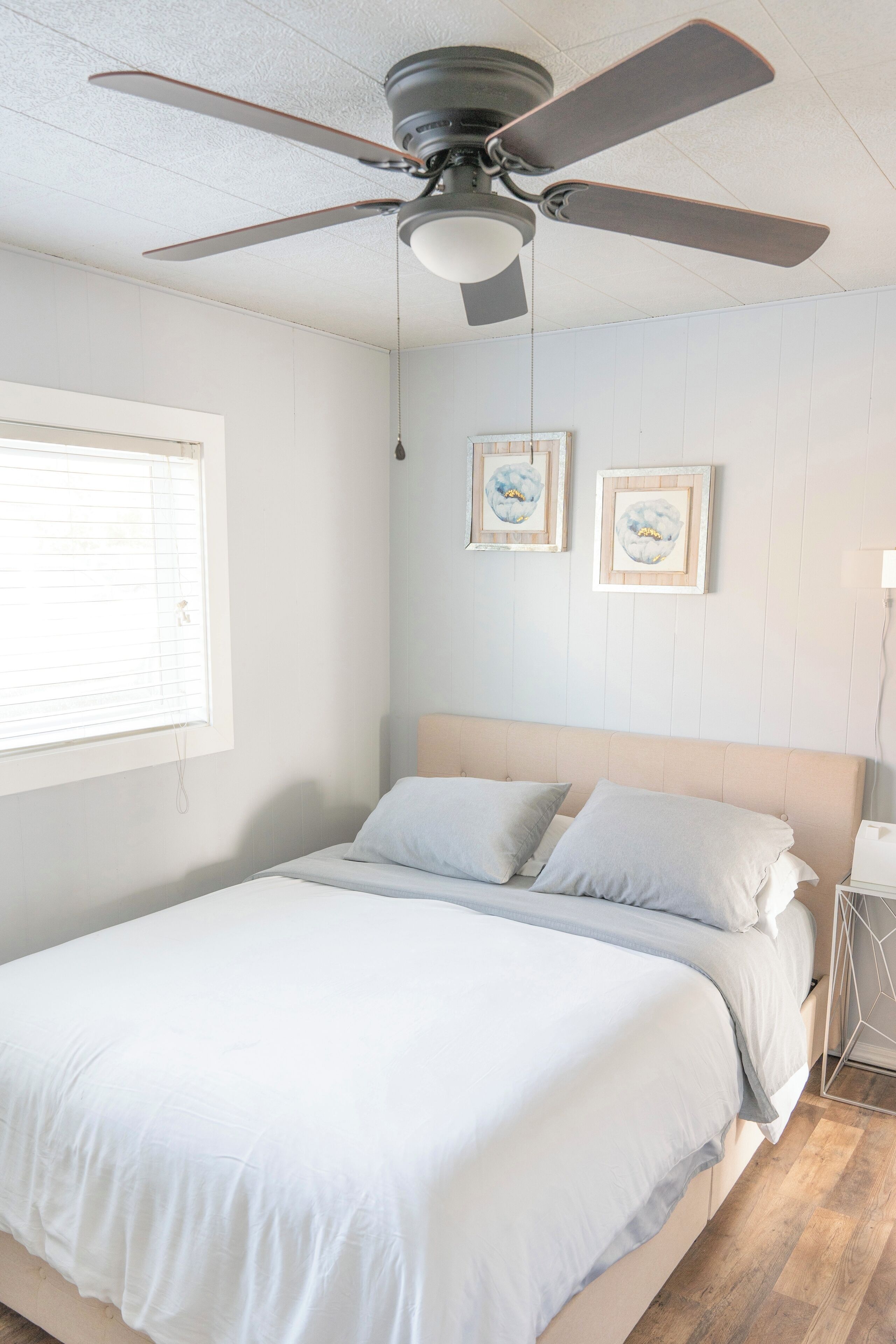 A bedroom with a bed, ceiling fan, window, and two framed pictures on the wall.