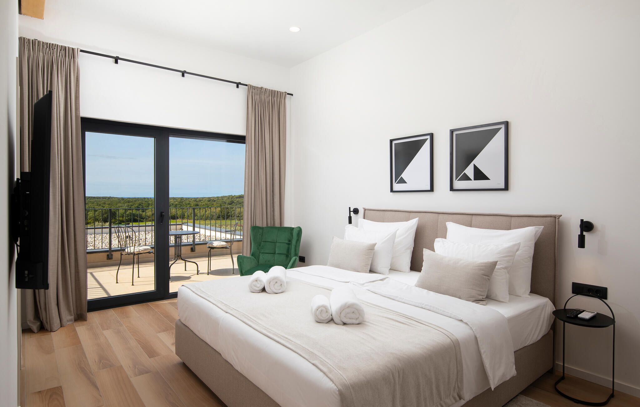 A hotel room with a large bed, a green chair, a balcony with a view, and two framed pictures on the wall.
