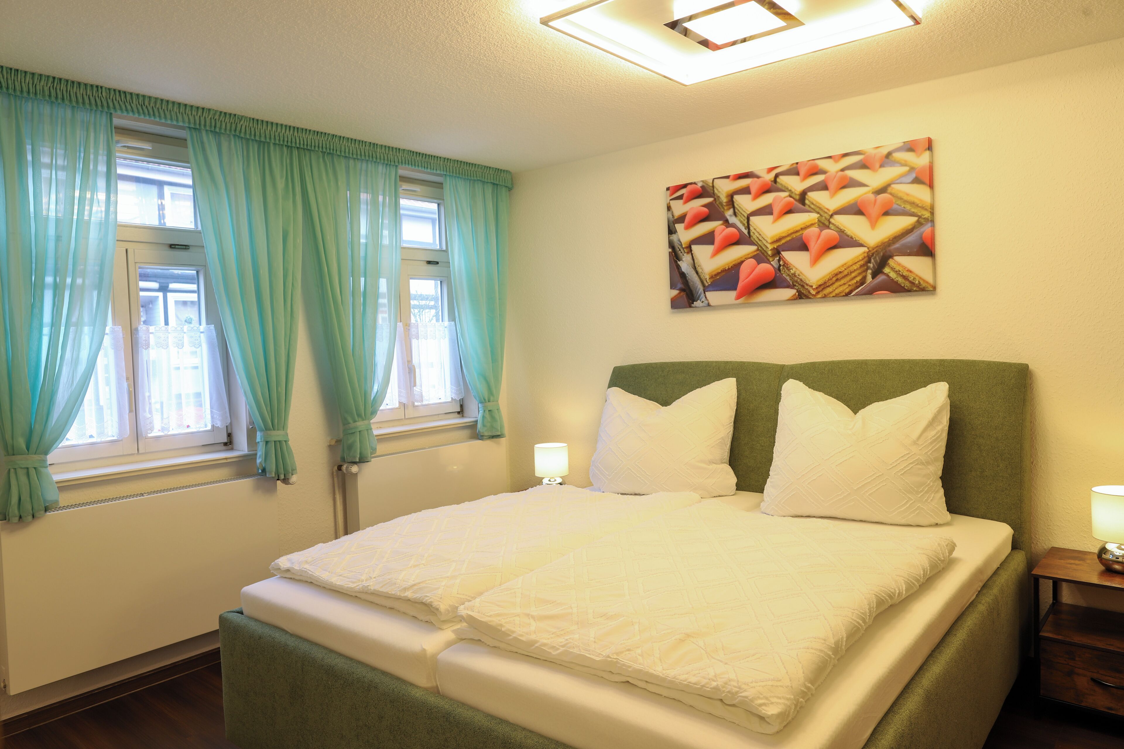 Un lit bien fait, avec du linge de lit blanc, une tête de lit verte et un tableau représentant des desserts accroché au mur.