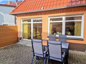 A patio with two chairs and a table, a house with orange walls and a red-tiled roof, and a window with curtains.