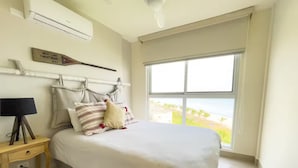 A bedroom with a bed, a nightstand, a lamp, a wall-mounted air conditioner, a window with a view, and a plaque on the wall. - Pacific Breeze -V5-5D- 2 bed, 2 bath (Punta Chame)