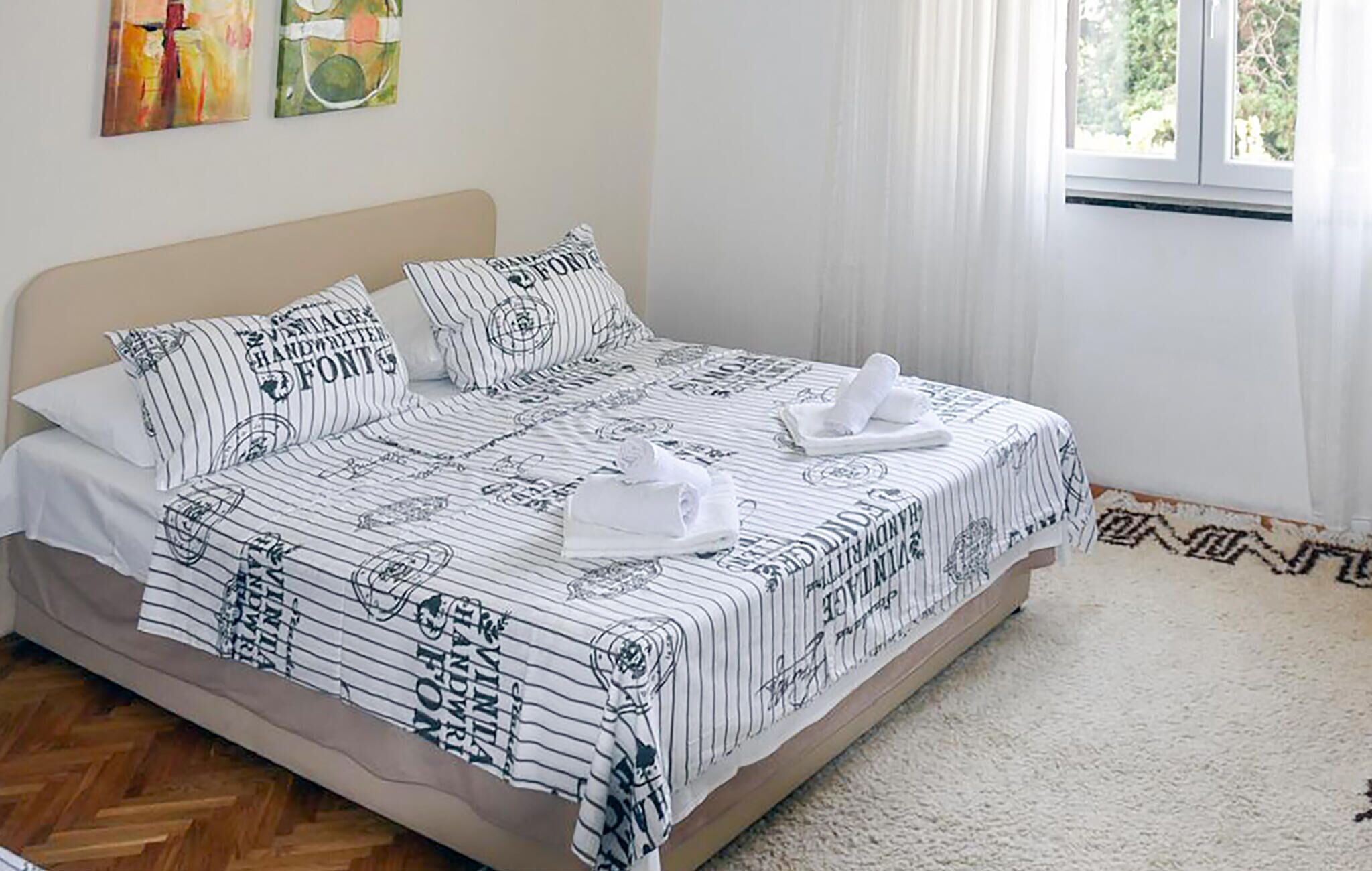 A neatly made bed with striped bedding and pillows, a window with curtains, and a herringbone-patterned carpet.