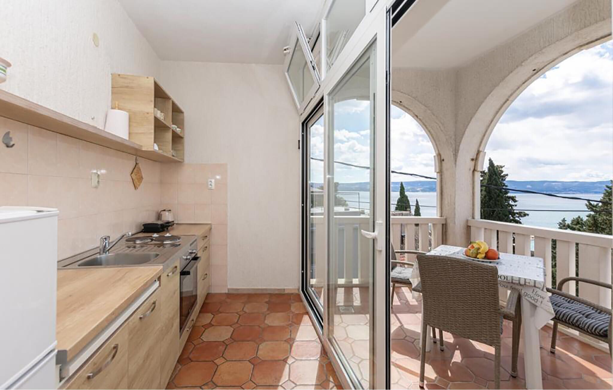 A balcony with a view of a lake and mountains, a small kitchen with a stove and sink, and a table with a chair.