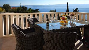 En balkon med fletstole og et spisebord med dække, der ud til et smukt havlandskab og fjerne bjerge.