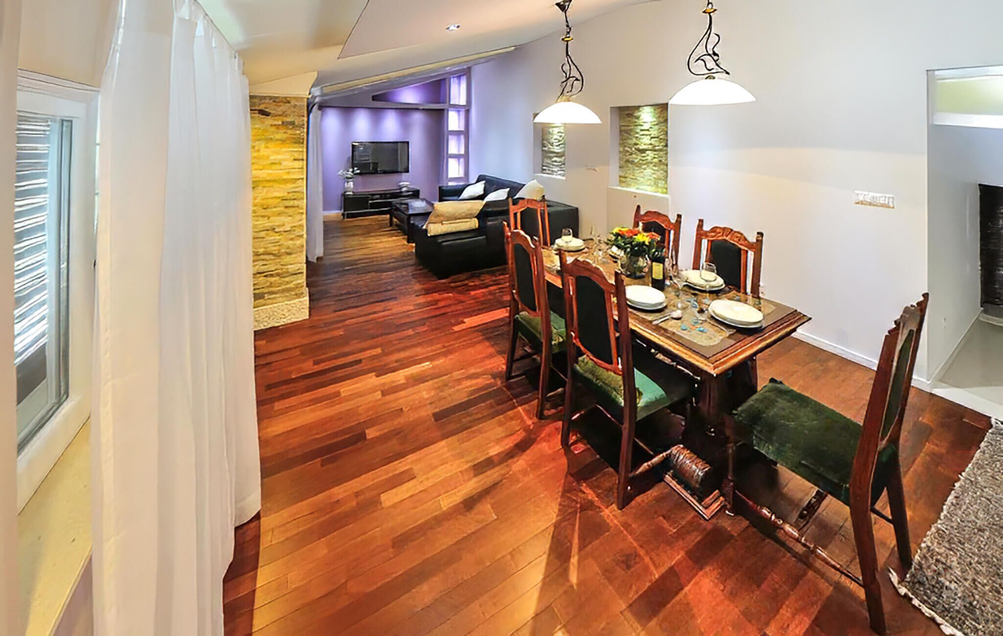 A dining area with a wooden table set for four, surrounded by chairs. A television is mounted on the wall in the background.