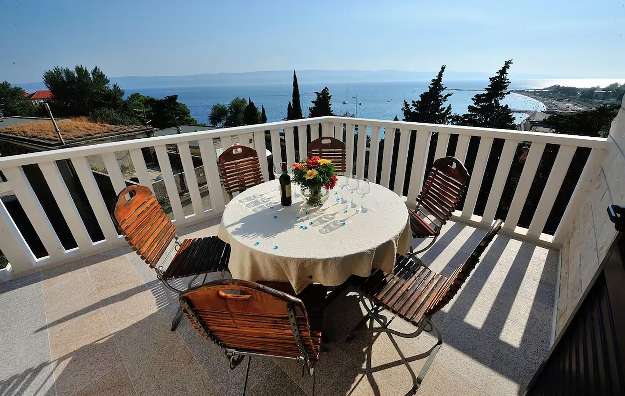 A round table with a white tablecloth, surrounded by wooden chairs, set on a balcony with a white railing.