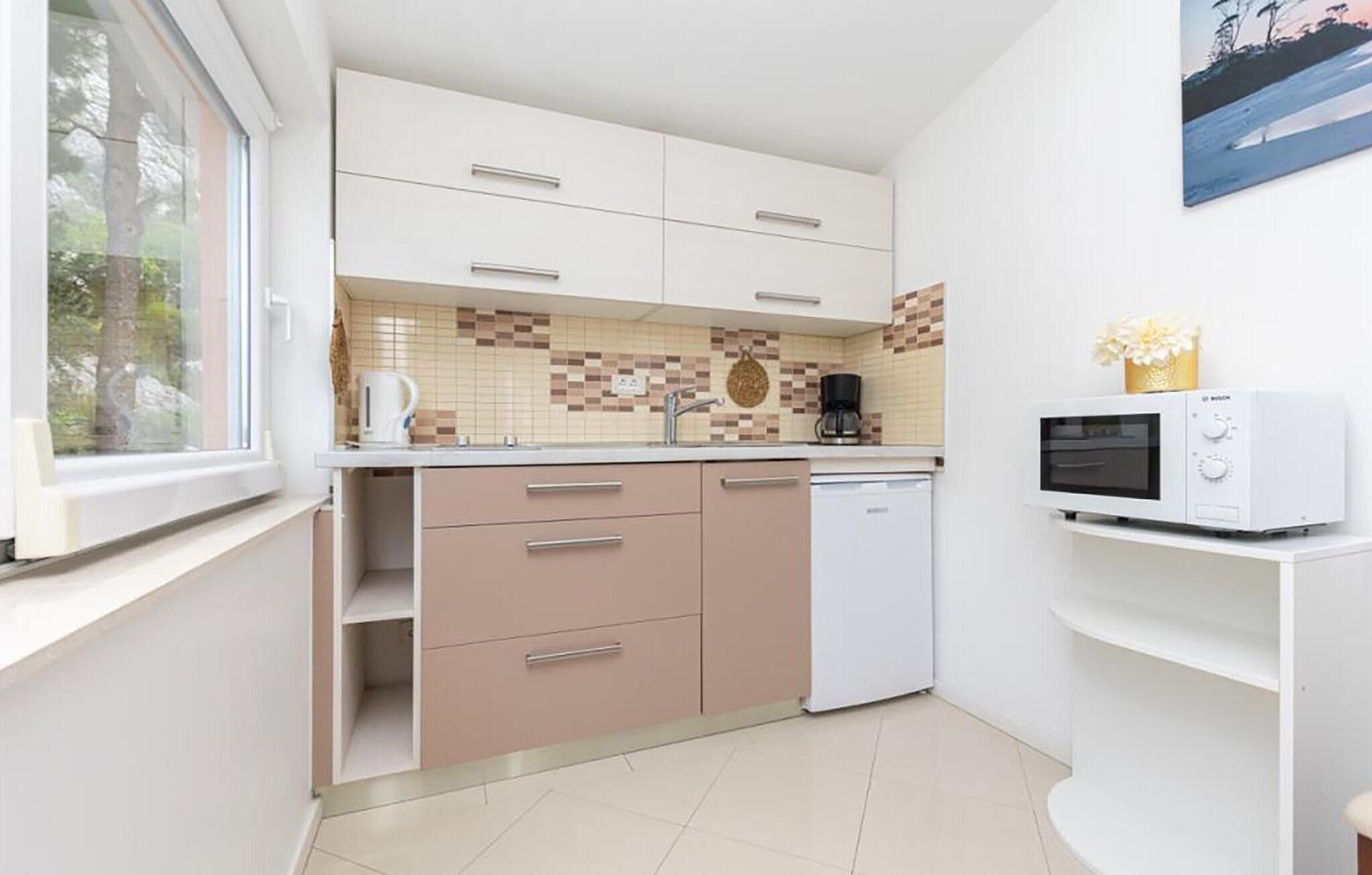 Una cocina compacta con armarios blancos, salpicadero de azulejos, cafetera, microondas y ventana con vistas a árboles.