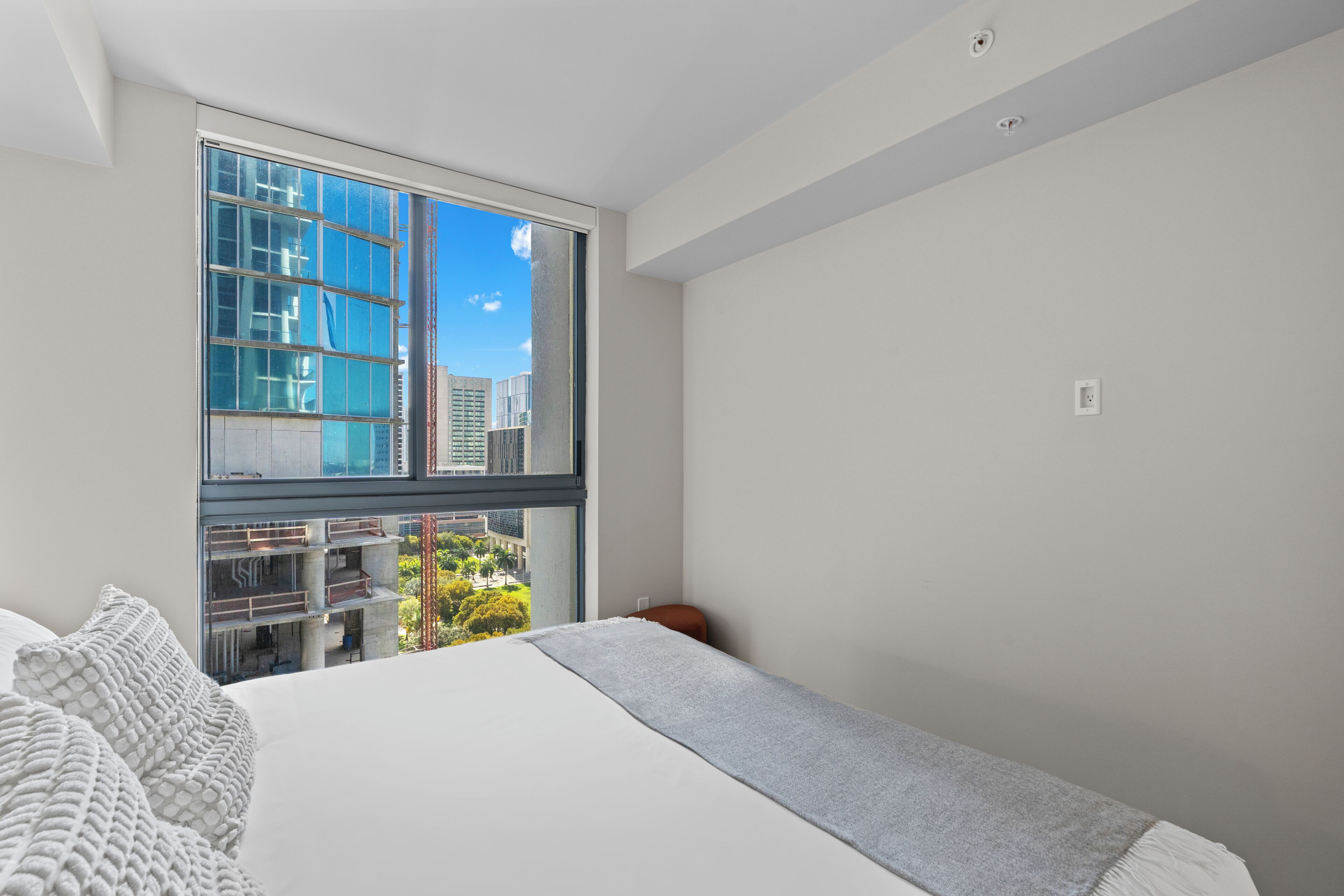 A modern bedroom with a large window, a bed with a grey blanket, and a view of a cityscape.