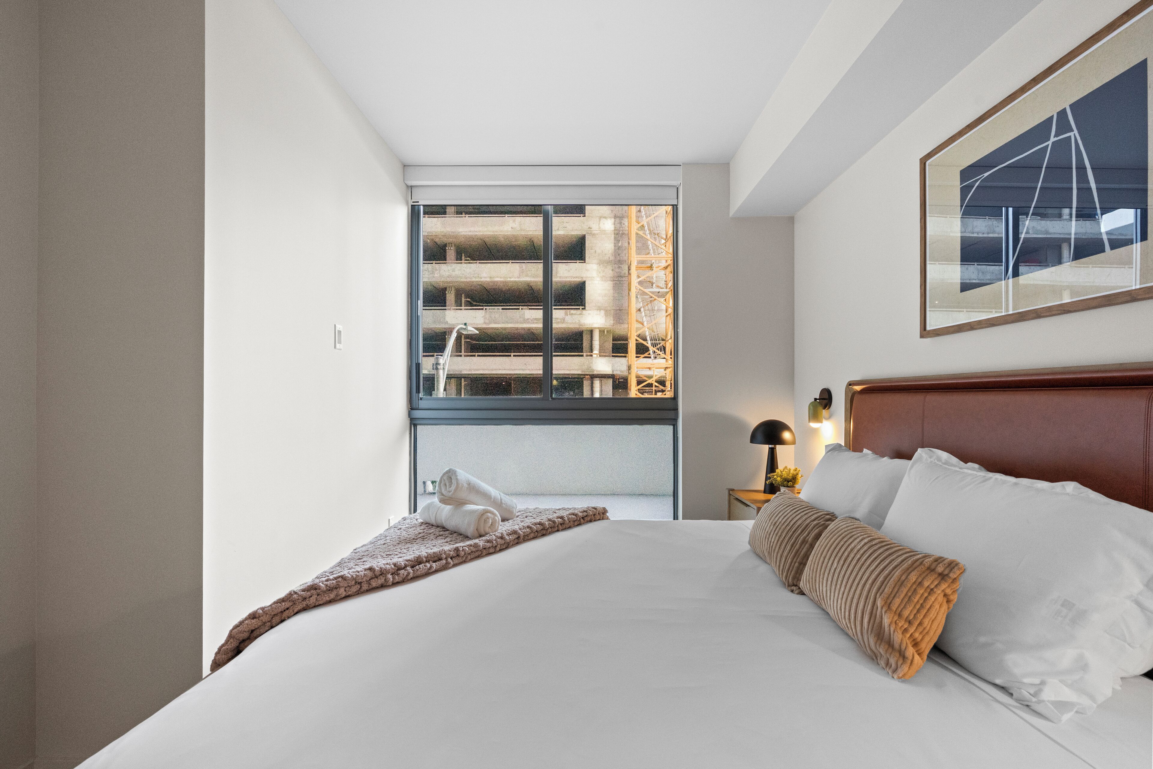 Una habitació d'hotel amb un llit gran, una finestra amb vista a un edifici en construcció, un quadre al mur i una taula de nit amb una làmpada.