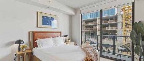 A modern hotel room with a large bed, a bedside table with a lamp, a framed picture on the wall, and a view of a construction site through the window.