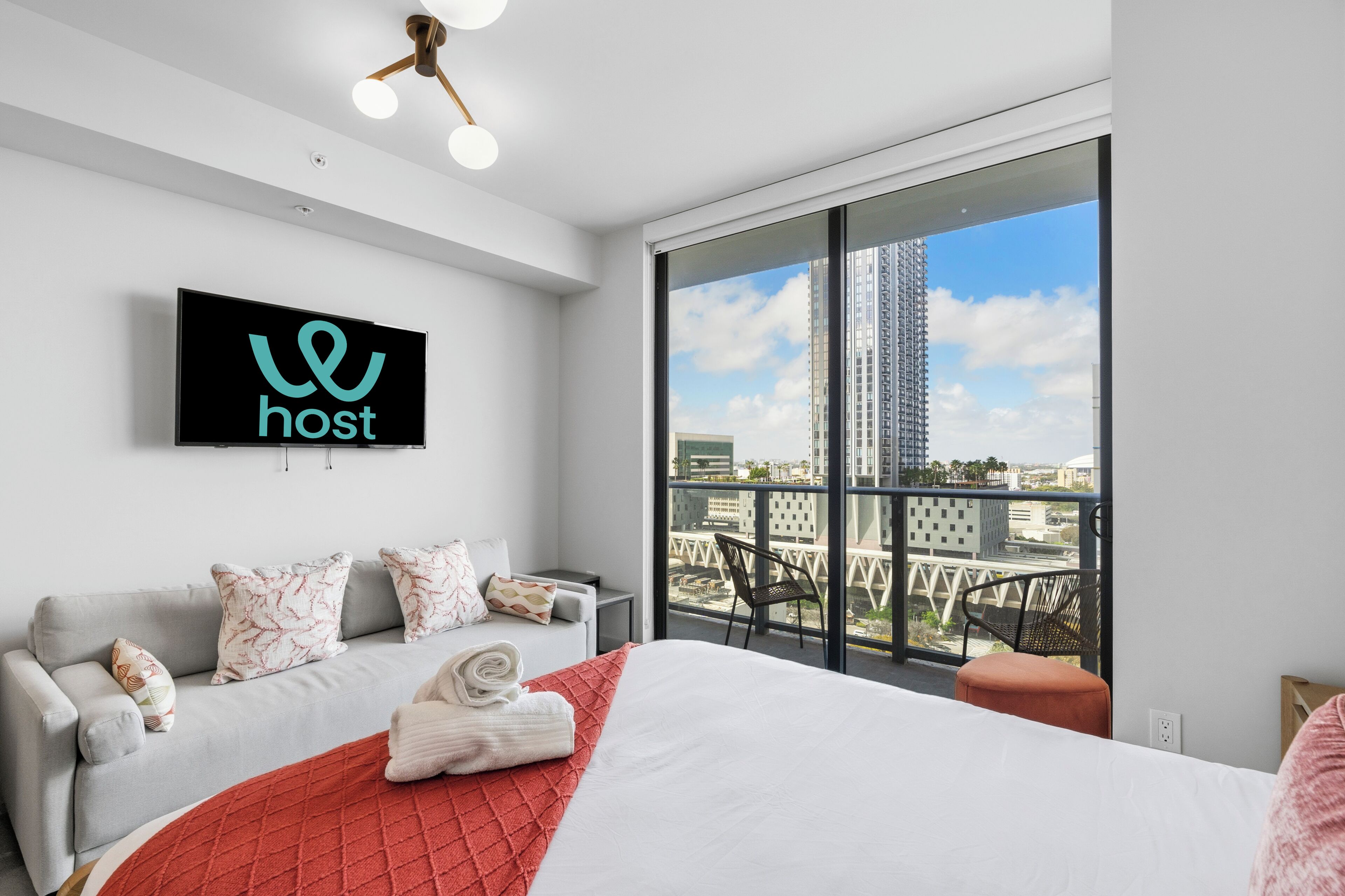 Ein modernes Hotelzimmer mit einem großen Bett, einem grauen Sofa, einem an der Wand montierten Fernseher und einem Balkon mit Blick auf die Stadt.