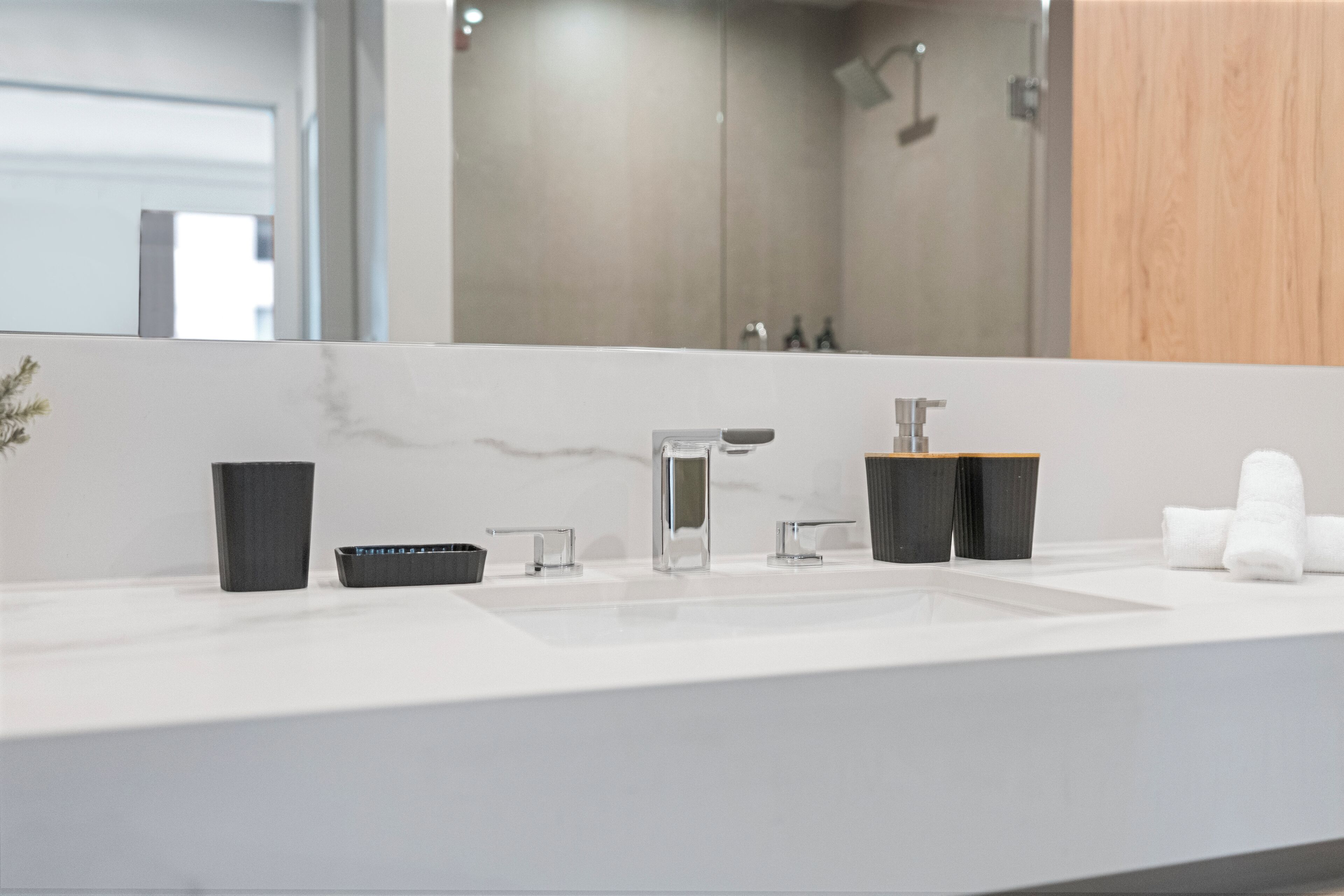 Een moderne badkamer met een grote spiegel, een wit wastafel, twee zwarte zeepdispensers en een houten kastje.
