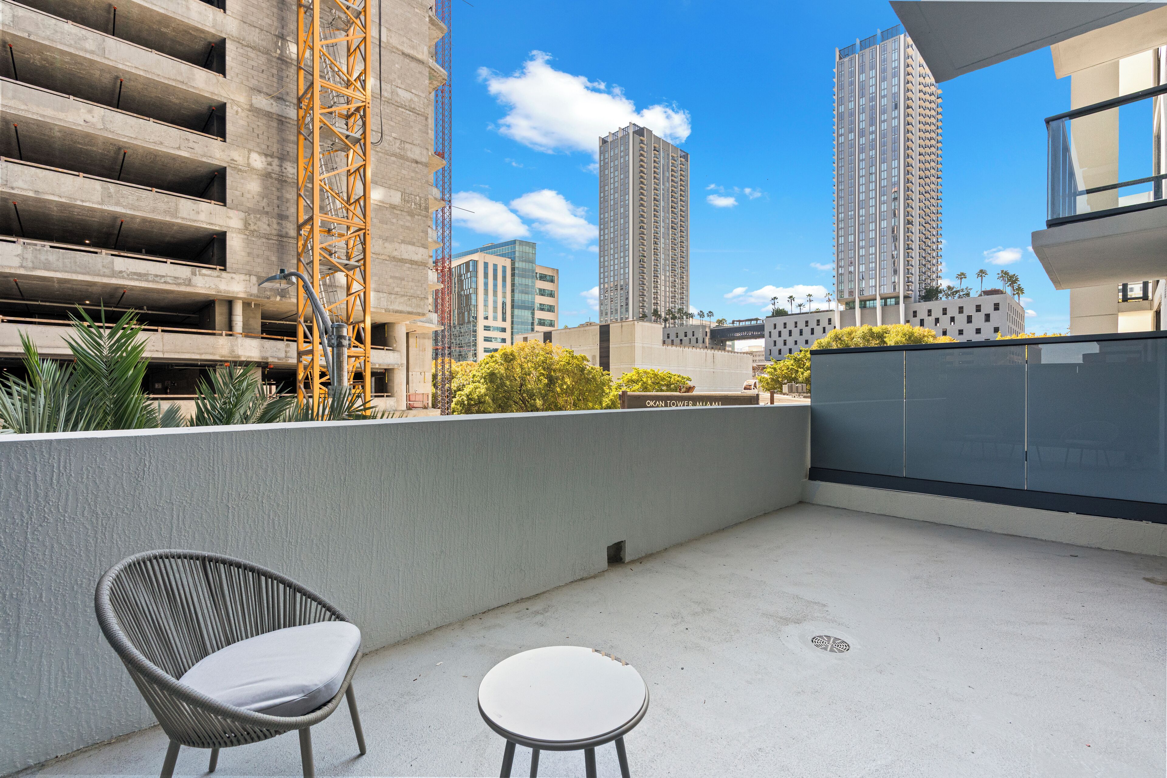 Un balcon avec une chaise en osier et une petite table, d’où l’on a vue sur un chantier de construction et de grands immeubles.