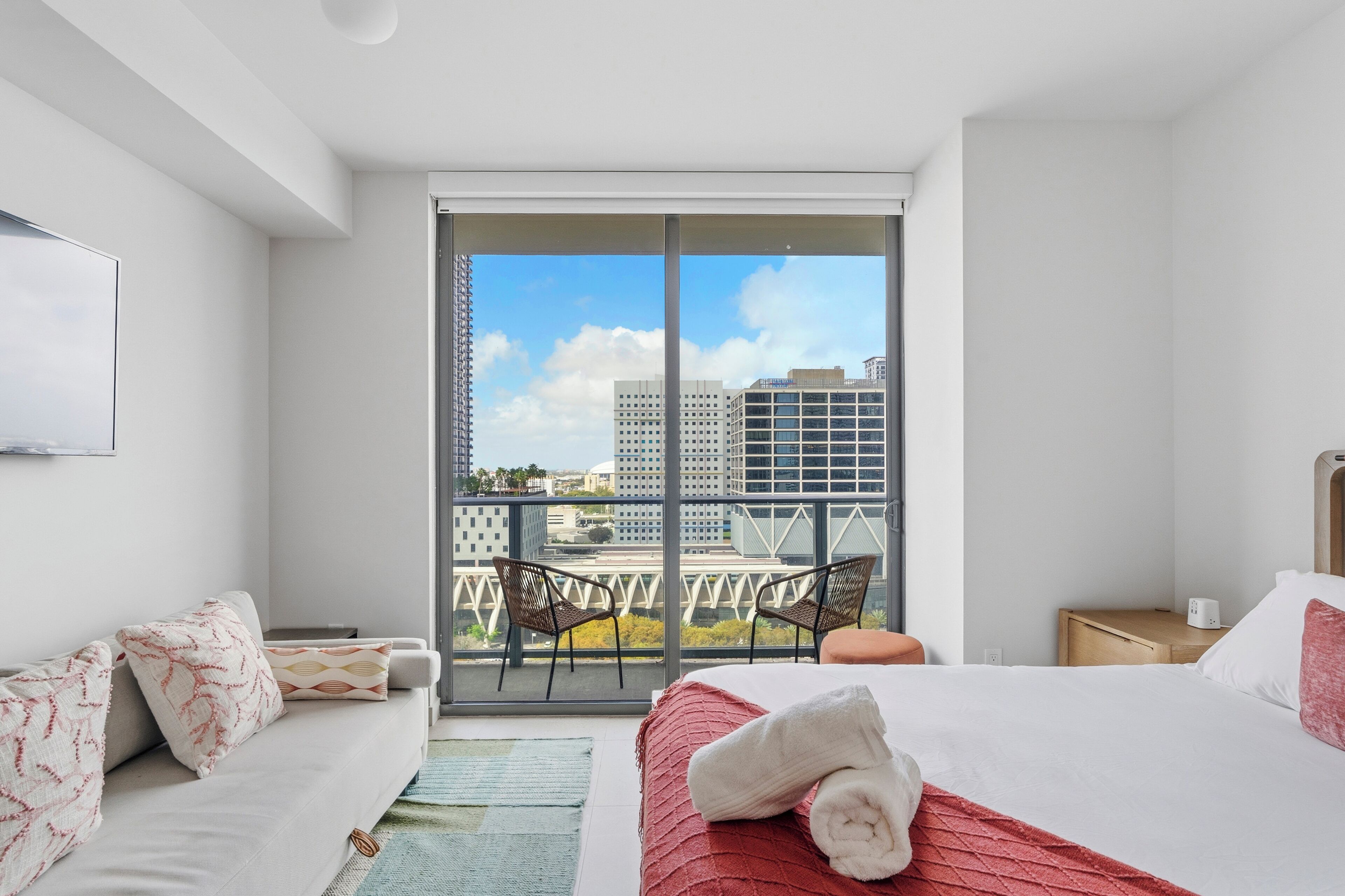 Een moderne hotelkamer met een groot balkon, een bed met een rode sprei, een bank met opvallende kussens en uitzicht op de stad.