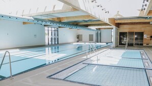 Une grande piscine intérieure dotée de plusieurs couloirs, d’un plafond en bois et d’une entrée avec porte en verre.