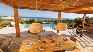 Ein Essbereich im Freien mit einem Holztisch, gedeckt für vier Personen, mit Blick auf einen Pool und eine schöne Aussicht auf Meer und Stadt.