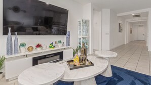 Ein modernes Wohnzimmer mit großem Flachbildfernseher, weißen Schränken, einem Marmortisch und einem blauen Teppich.
