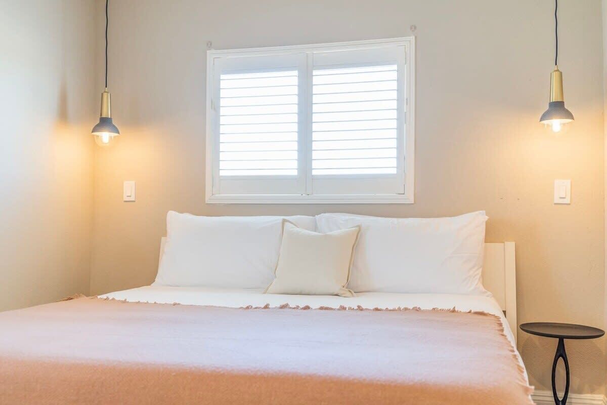A bedroom with a bed, white bedding, a window with blinds, and two pendant lights.