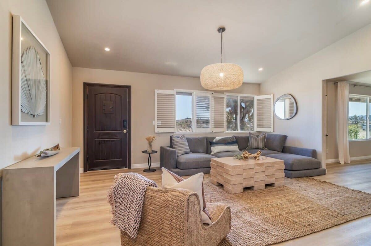 A spacious living room with a grey sofa, a coffee table, and a woven chair. There is a large window with shutters, a dark wooden door, and a hanging light fixture.