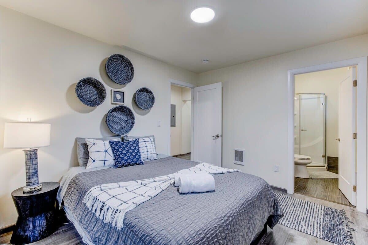 Un dormitorio ordenado con una cama grande, una mesita de noche, una lámpara de pared y una puerta que da a un baño.