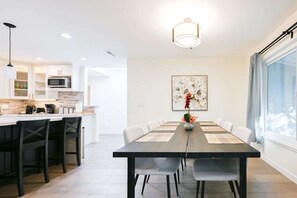 A modern kitchen with white cabinets, a microwave, and a coffee machine. There is a dining area with a long table and chairs, and a large window with curtains. - Marbella Lane - Exquisite Home 3BR (Sunnyvale)