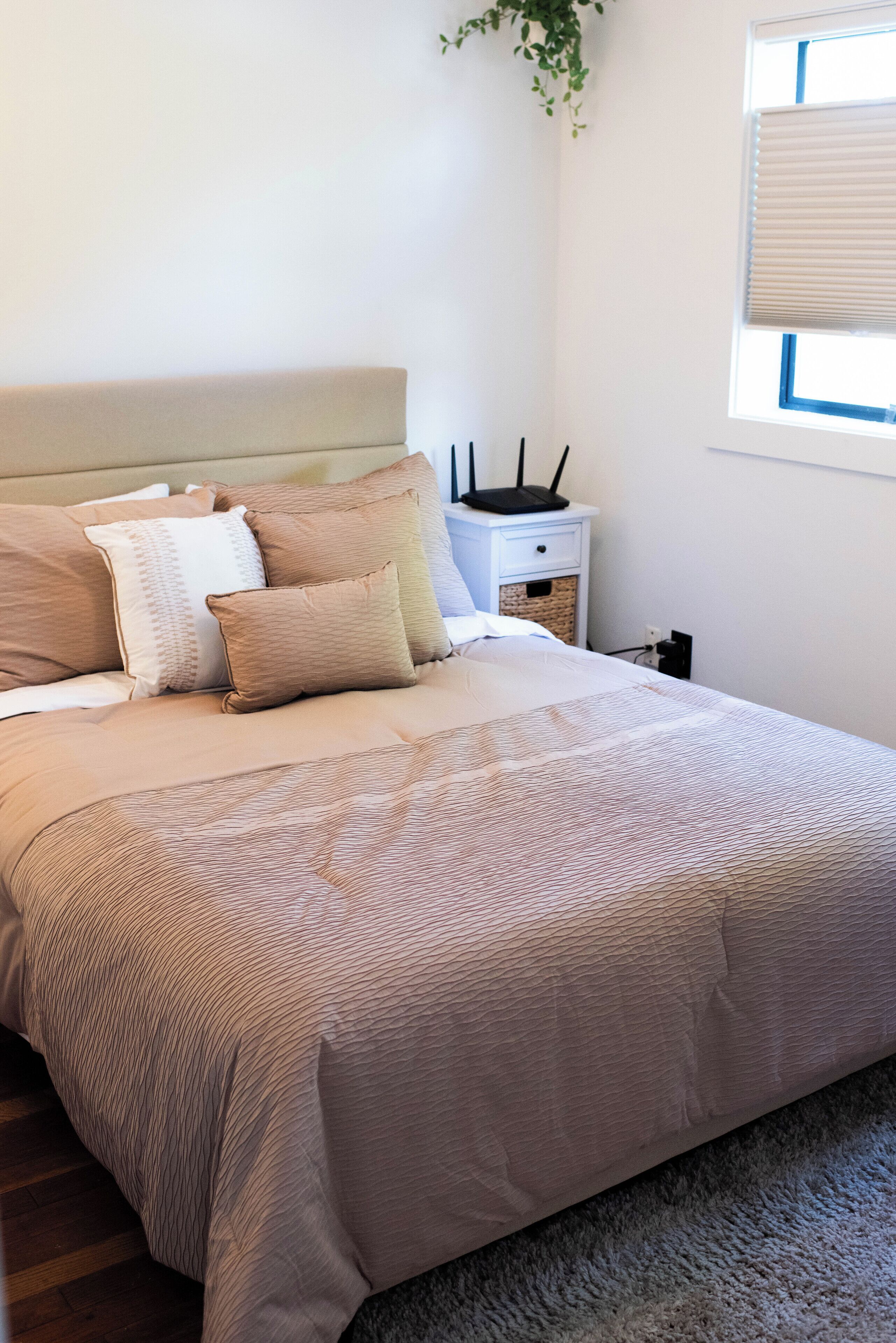 A neatly made bed with a beige comforter and pillows, a white bedside table with a lamp, and a window with blinds.