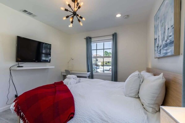 A bedroom with a bed, a TV mounted on the wall, a window with curtains, and a painting on the wall.
