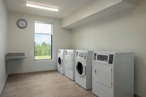 A laundry room with three white front-loading washers and dryers, a clock on the wall, and a window with a view of trees.
