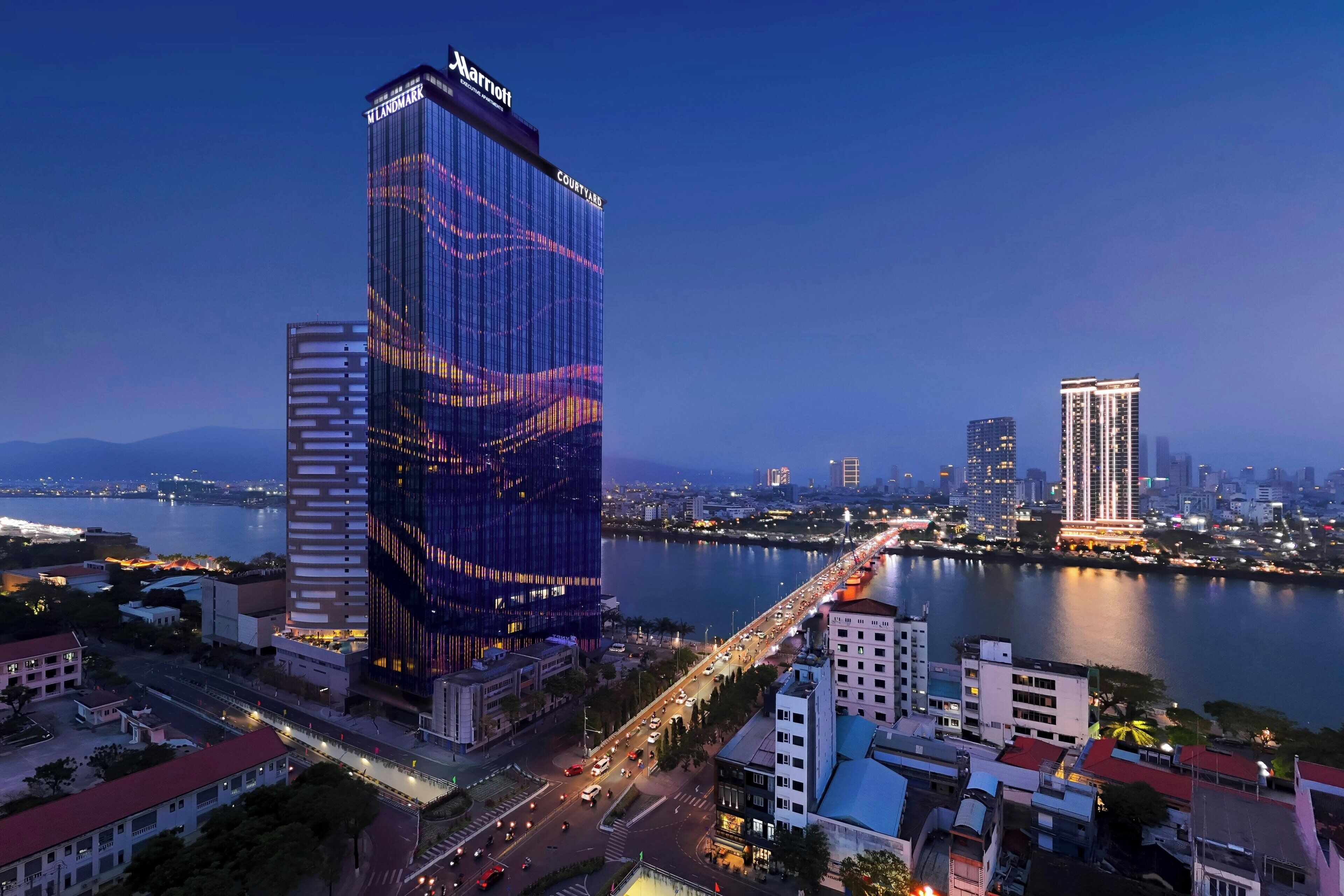 Um hotel moderno com fachada de vidro, um letreiro chamativo da 'Marriott' e vista para uma movimentada orla com uma ponte.