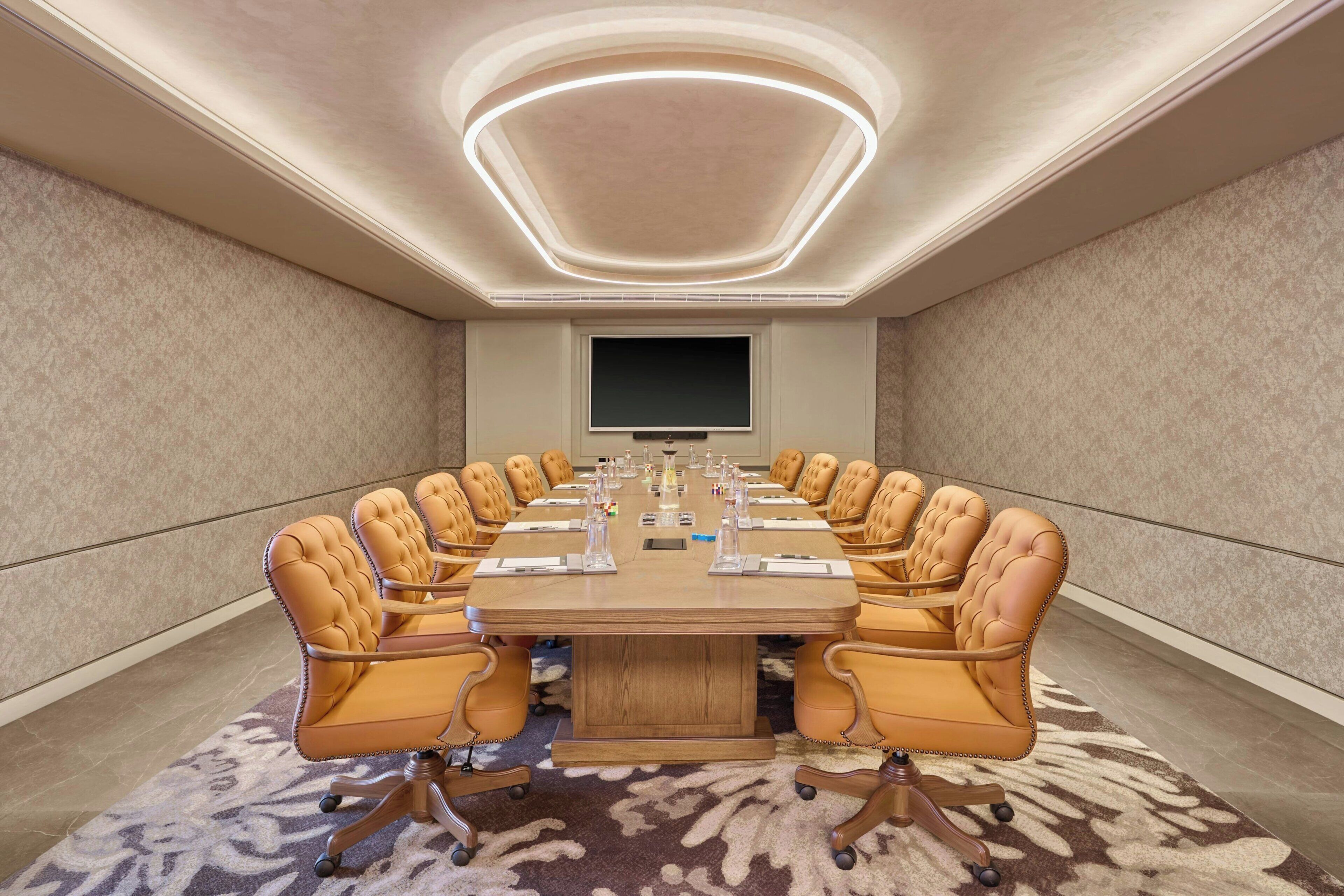 A conference room with a large oval table, leather chairs, a flat-screen TV, and a patterned carpet.