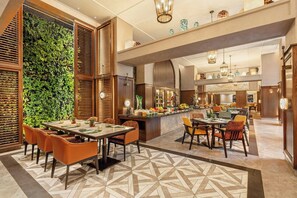 A modern hotel lobby with a dining area, a bar, and a kitchen visible in the background.