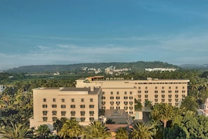 A hotel with multiple floors, surrounded by palm trees and a hilly landscape in the background.