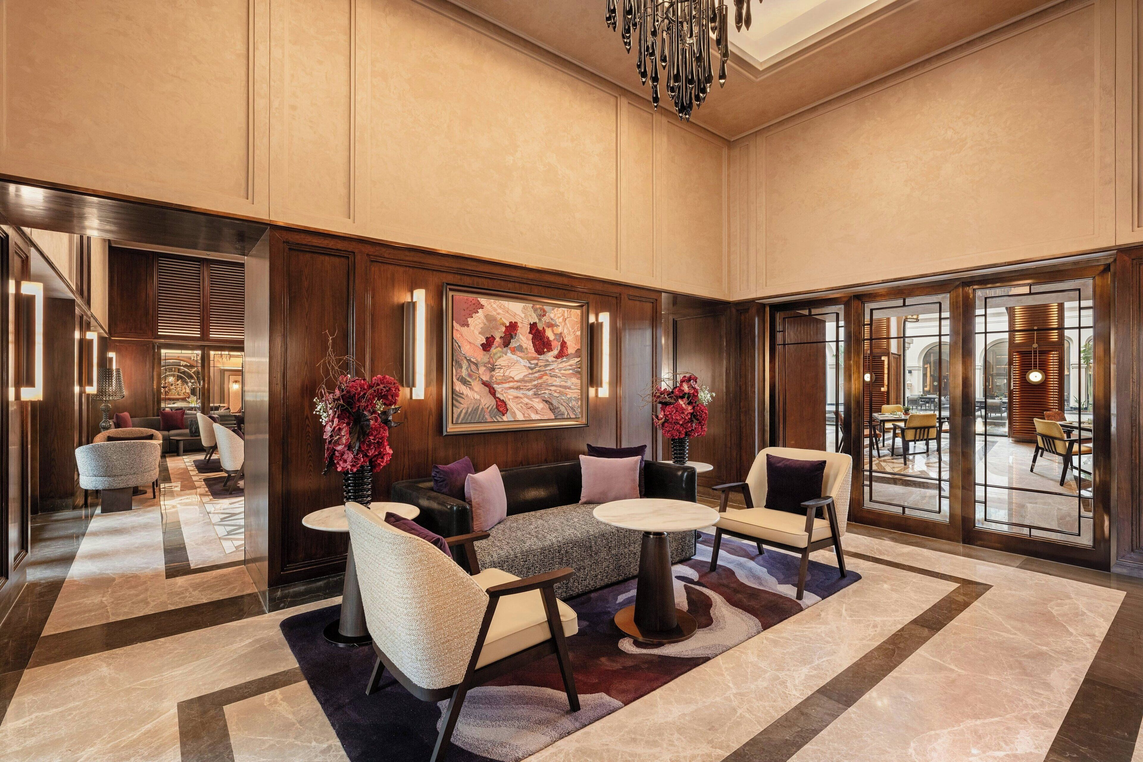 Eine großzügige Lobby mit einer Couch, Sesseln und einem Couchtisch. Große Fenster und ein Kronleuchter sind ebenfalls vorhanden.