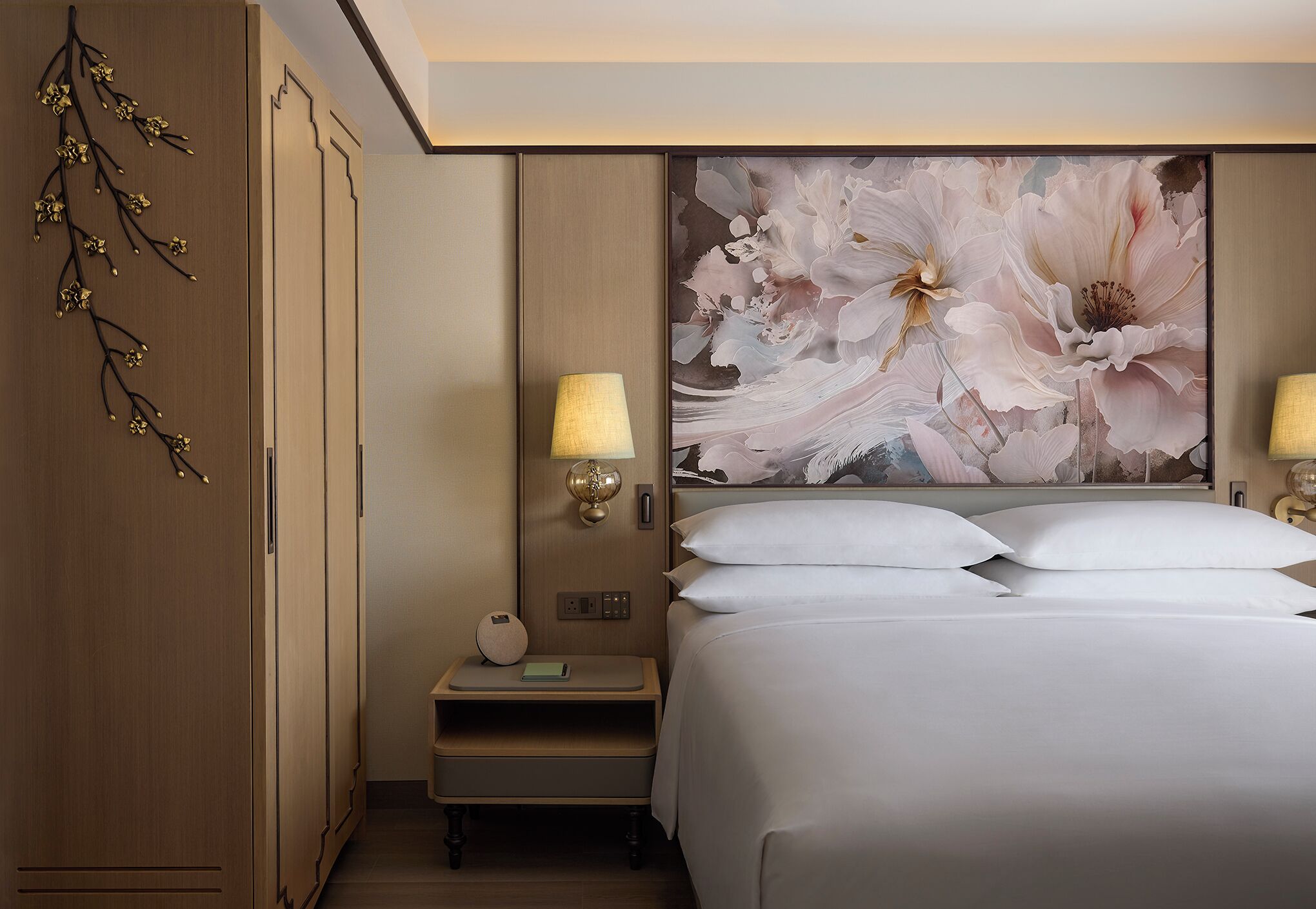 A hotel room with a large bed, a nightstand with a lamp, a wall-mounted lamp, and a decorative branch on the left wall.
