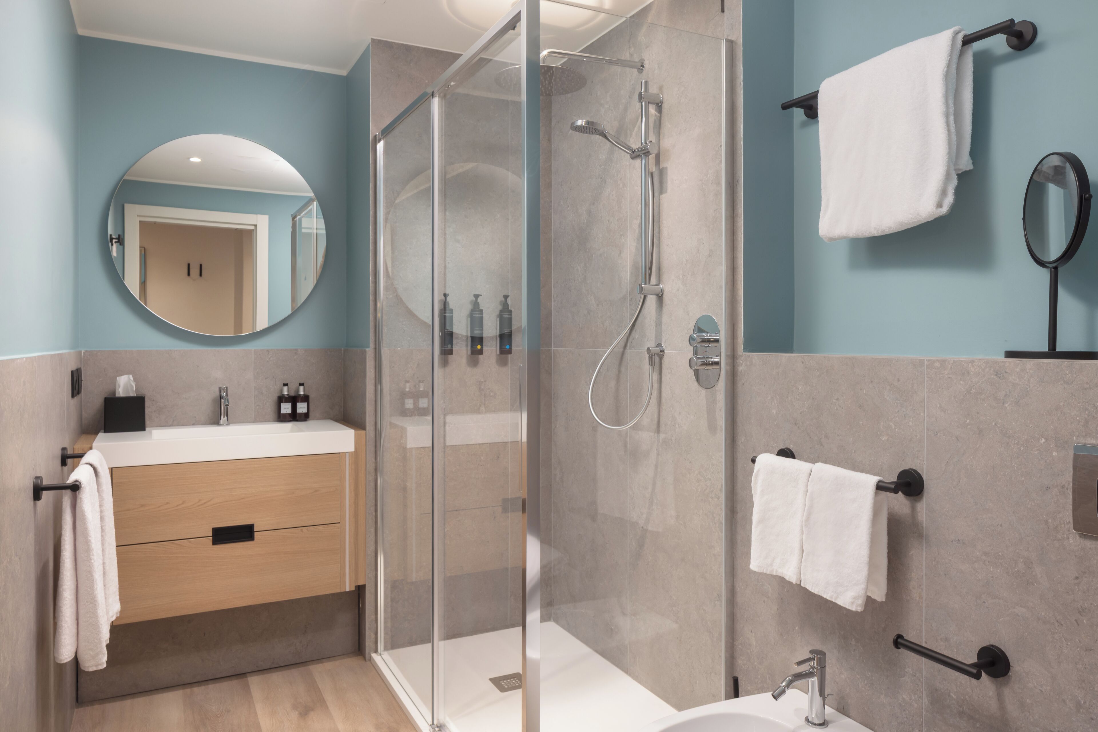 A modern bathroom with a glass-enclosed shower, a wooden vanity, a round mirror, and a towel rack.