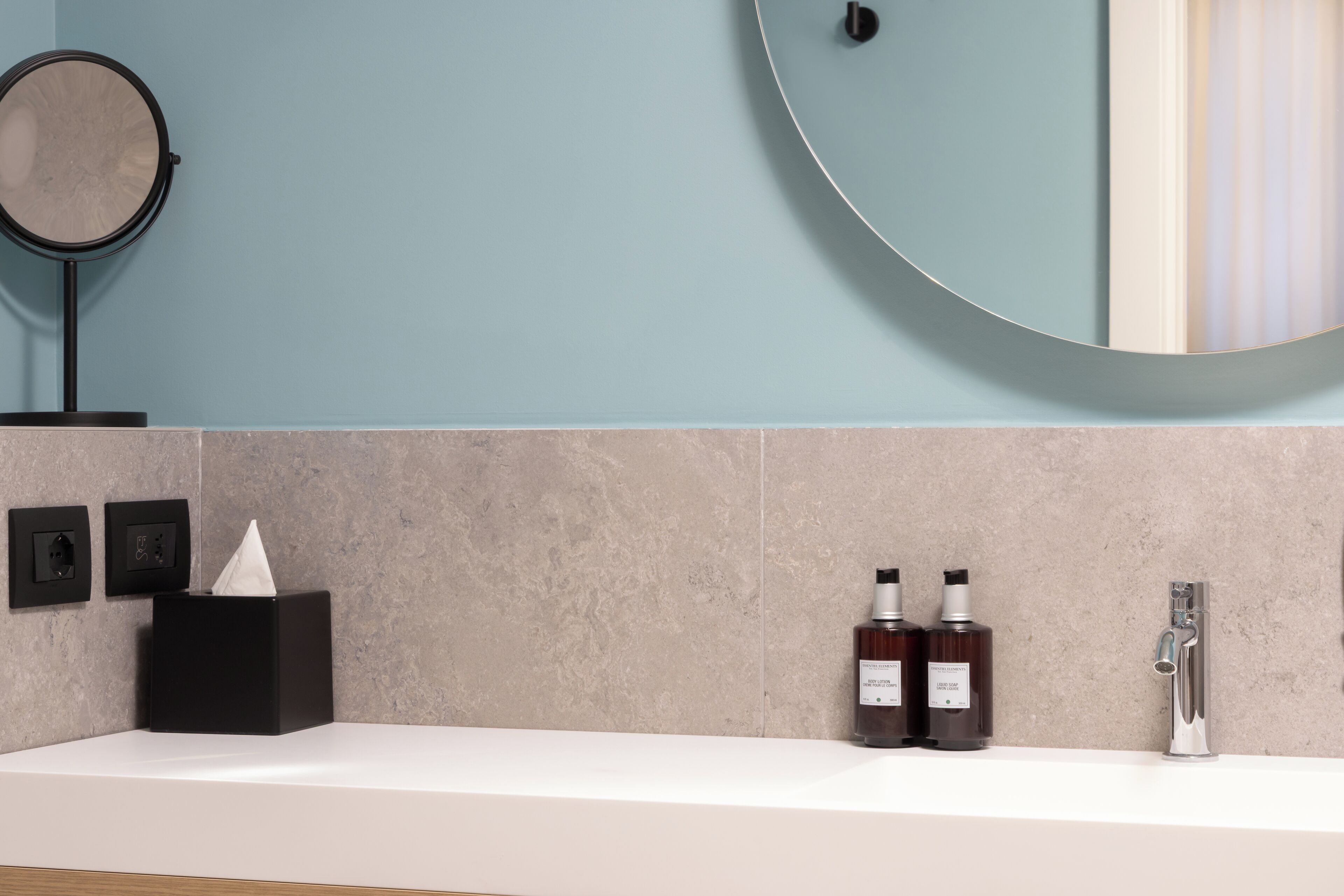 A bathroom with a white countertop, a round mirror, a black tissue box, two bottles of hand soap, and a modern faucet.