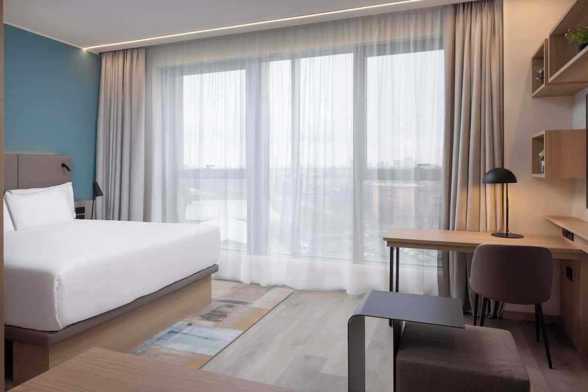 A modern hotel room with a large bed, a desk, a chair, and a view of the city through sheer curtains.